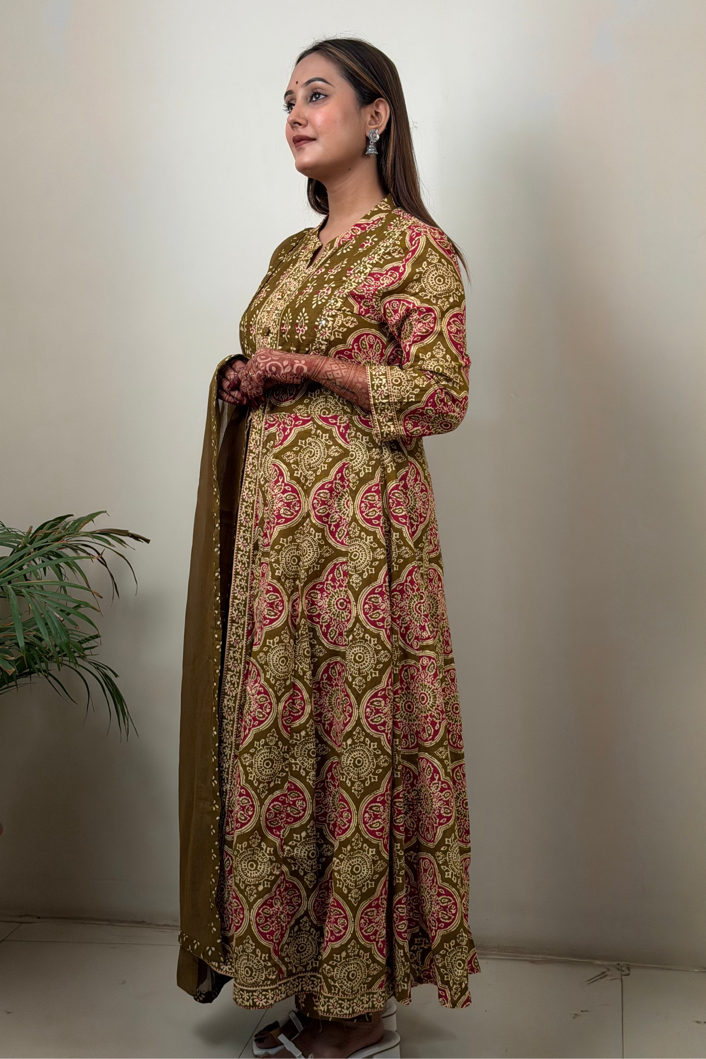 Woman wearing a patterned dress holding a dupatta against a plain wall.