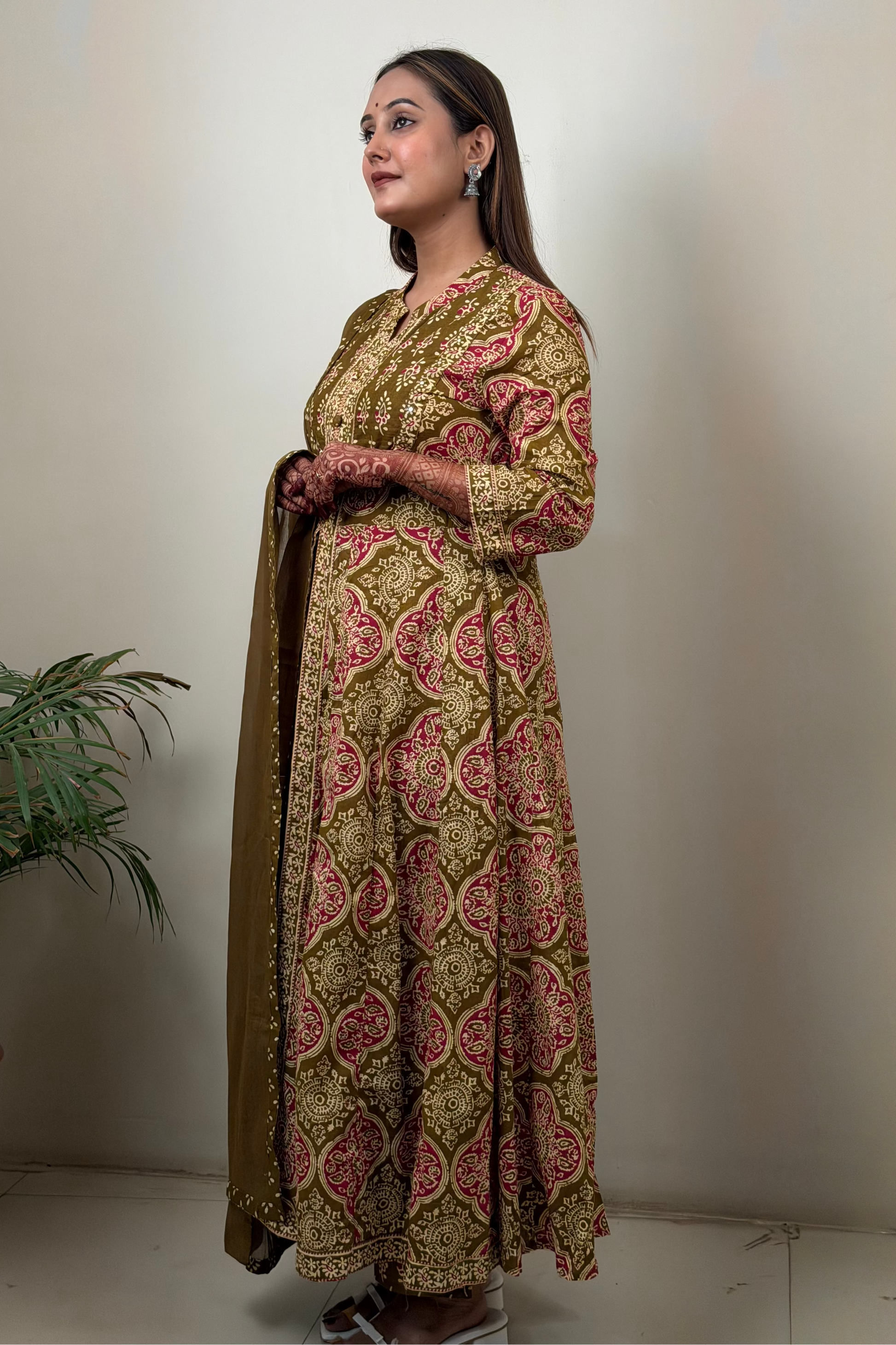 Woman wearing a patterned dress holding a dupatta against a plain wall.