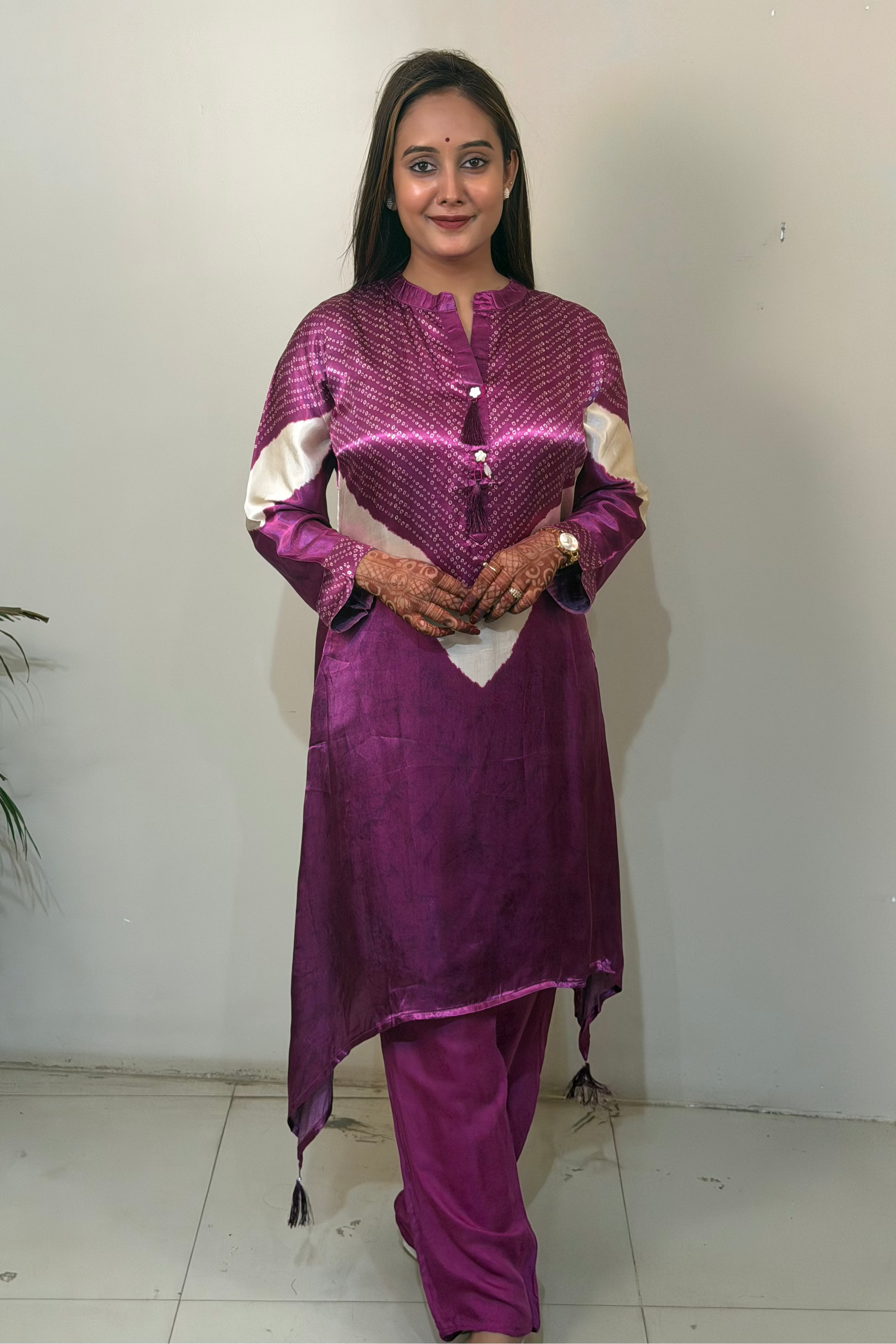 Woman wearing a purple traditional outfit with white accents against a plain wall.