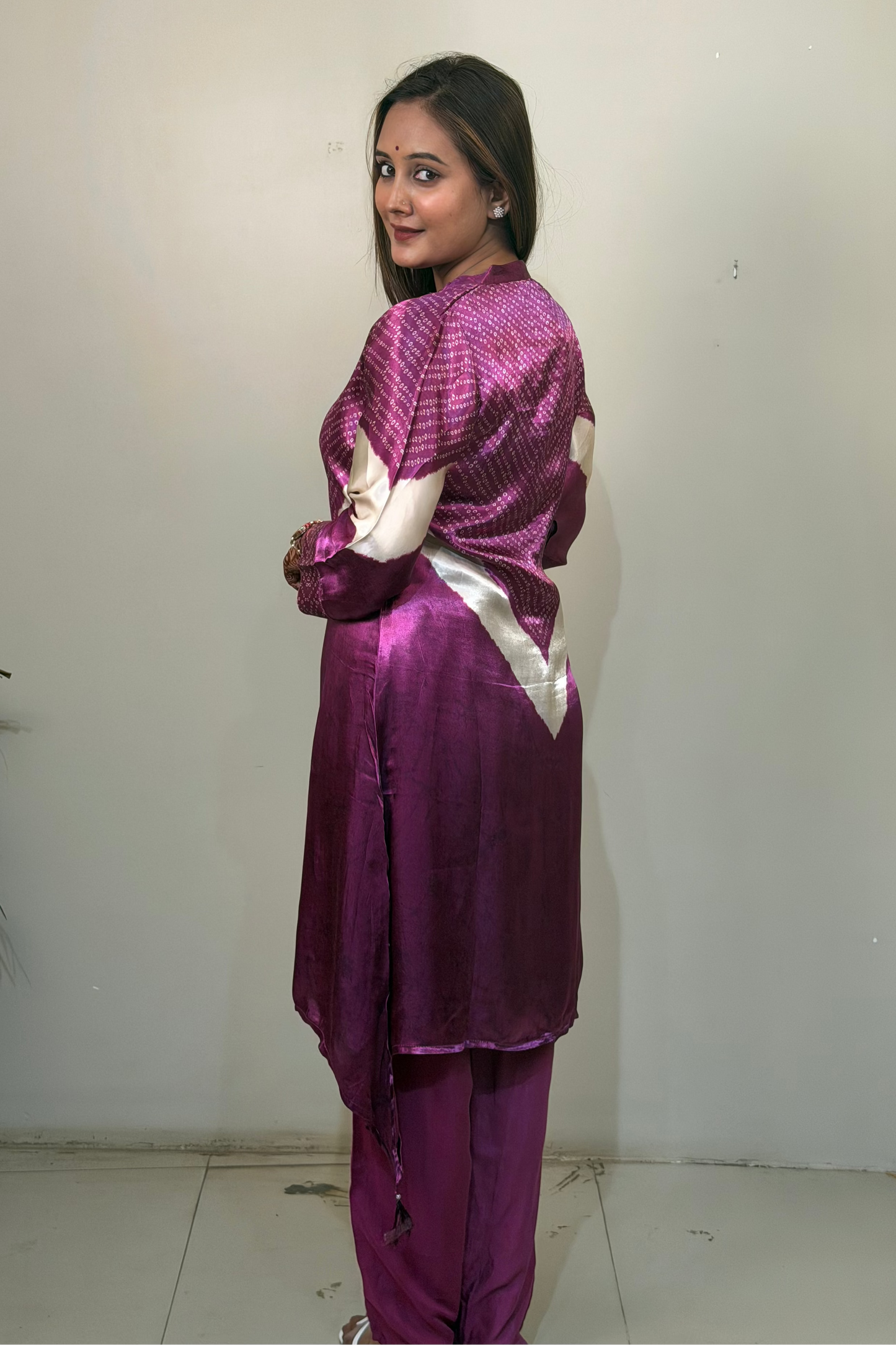 Woman wearing a purple traditional outfit with white accents against a plain wall.